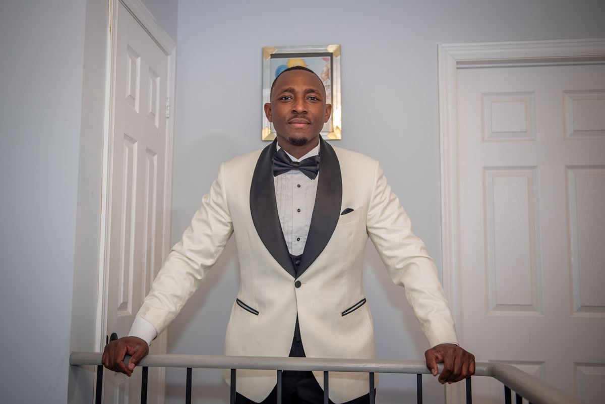 A man in a white tuxedo stands on a staircase railing, looking at the camera, with two white doors behind him.