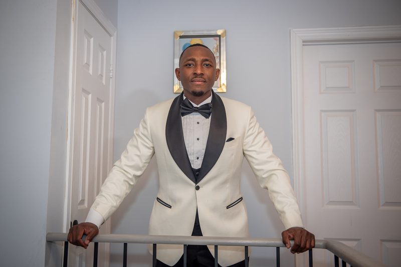 A man in a white tuxedo stands on a staircase railing, looking at the camera, with two white doors behind him.