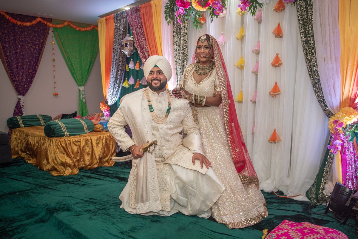 A smiling South Asian bride and groom in traditional attire pose beside each other in an ornate setting.