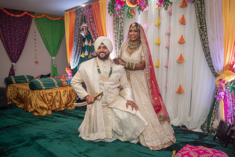 A smiling South Asian bride and groom in traditional attire pose beside each other in an ornate setting.