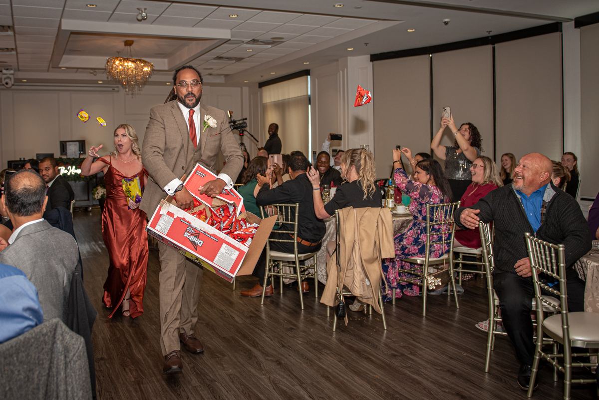 A groom in a brown suit walks through a cheering crowd at a wedding reception, holding an open box of Doritos.