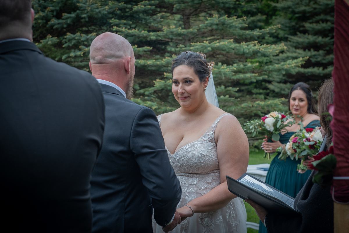 Bride in lace dress smiles at groom, holding hands near bridesmaids and officiant during outdoor ceremony.
