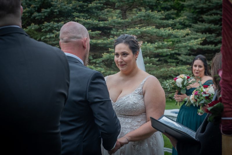 Bride in lace dress smiles at groom, holding hands near bridesmaids and officiant during outdoor ceremony.