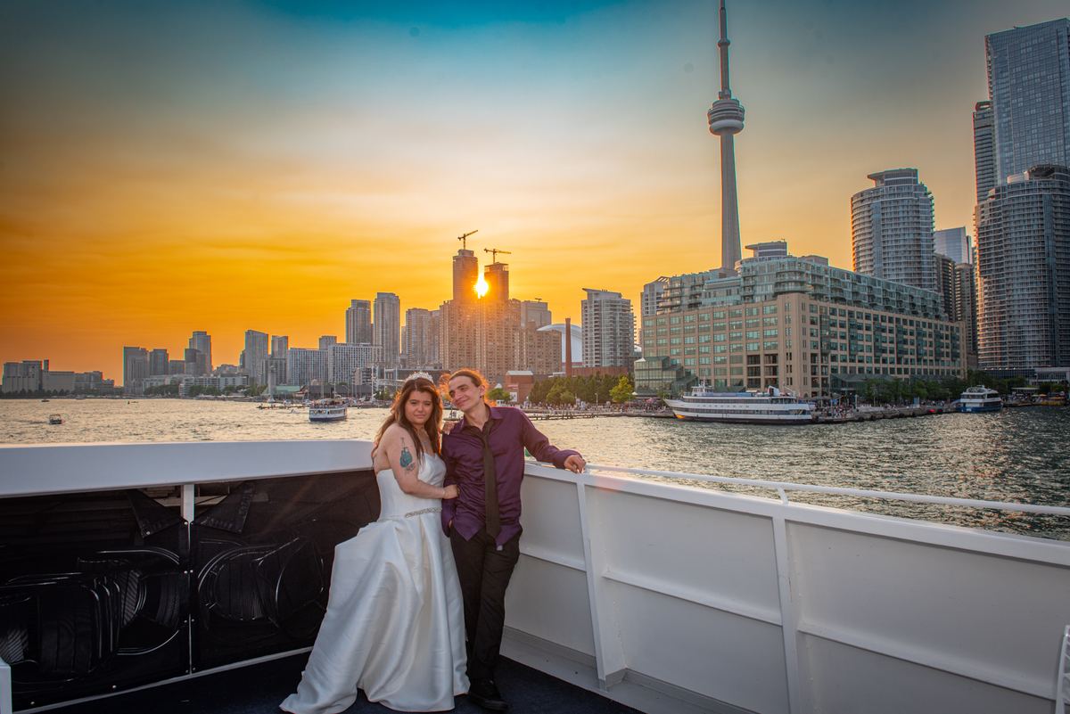 Ashley and Nicholas pose on the deck of a boat during a vibrant sunset, with Toronto's skyline glowing in the background.