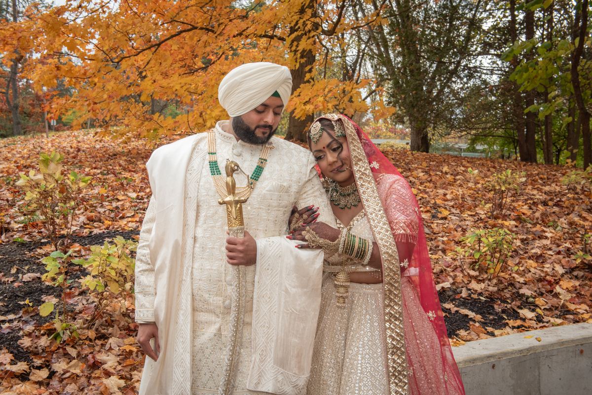 A Sikh groom in white and bride in red gold lehenga stand close in autumn park, leaves carpet ground.