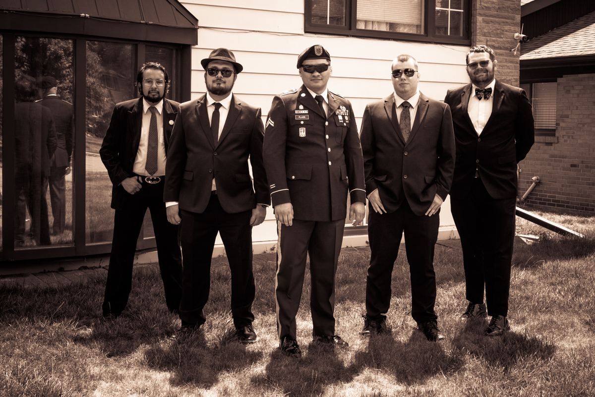 Five formally dressed men, including a soldier in uniform, pose outdoors in front of a house during the late afternoon.