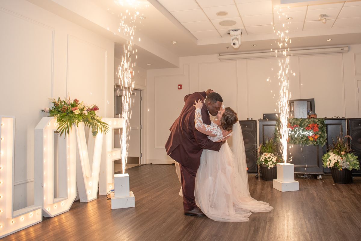A bride and groom kiss during their first dance, surrounded by fireworks and floral arrangements in a warmly lit hall.
