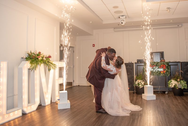 A bride and groom kiss during their first dance, surrounded by fireworks and floral arrangements in a warmly lit hall.