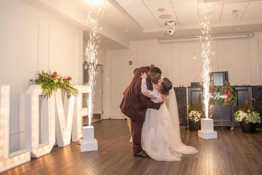 A bride and groom kiss during their first dance, surrounded by fireworks and floral arrangements in a warmly lit hall.