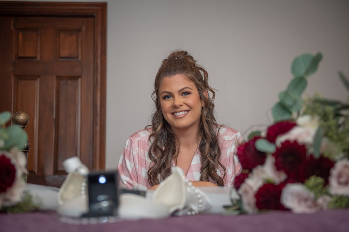 A woman smiles warmly while seated at a table adorned with flowers and a ring box, indicating an engagement moment.