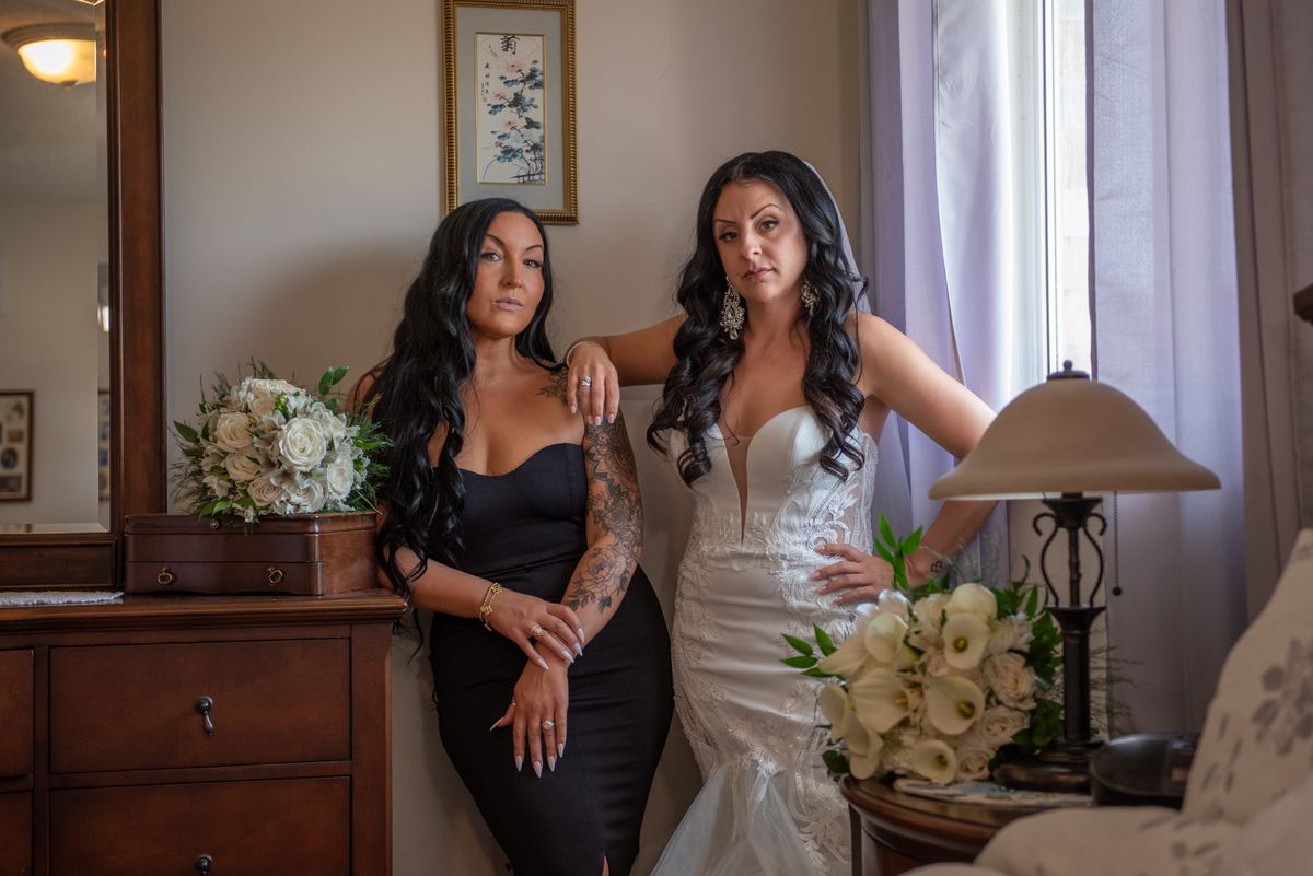 Two women in a warmly lit room, one in a wedding gown holding flowers, the other with a tattoo and ring.