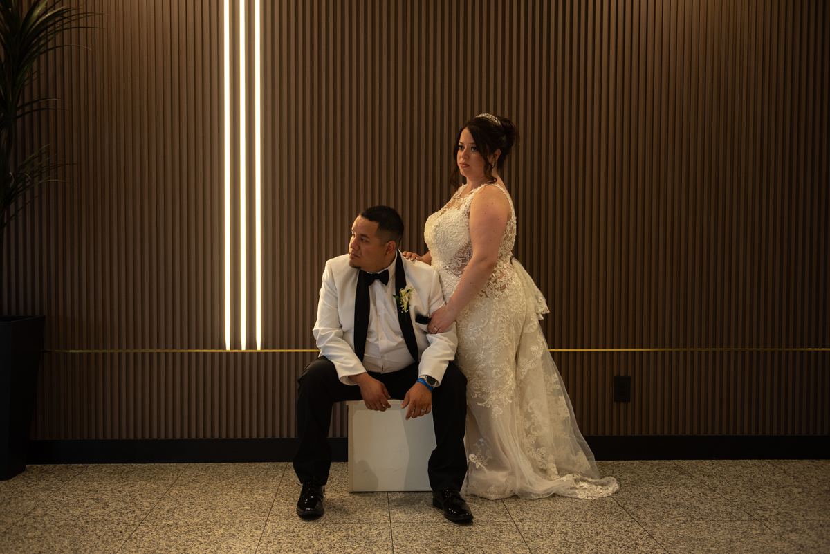 A newlywed couple poses tenderly in a modern, warmly lit room with soft golden light from wood-paneled walls.
