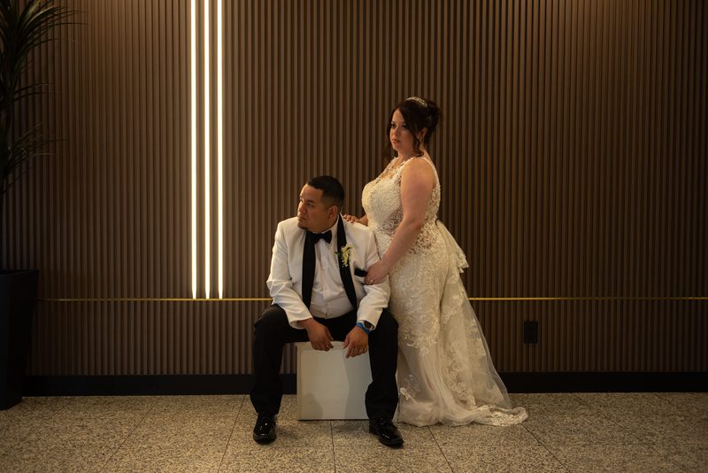 A newlywed couple poses tenderly in a modern, warmly lit room with soft golden light from wood-paneled walls.