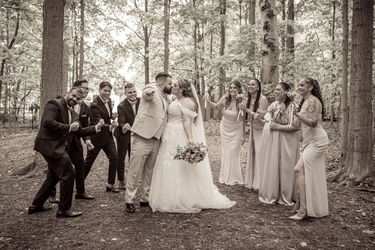 A bride and groom kiss in a wooded clearing, surrounded by their laughing wedding party pointing at the camera.