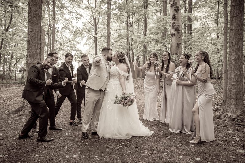 A bride and groom kiss in a wooded clearing, surrounded by their laughing wedding party pointing at the camera.