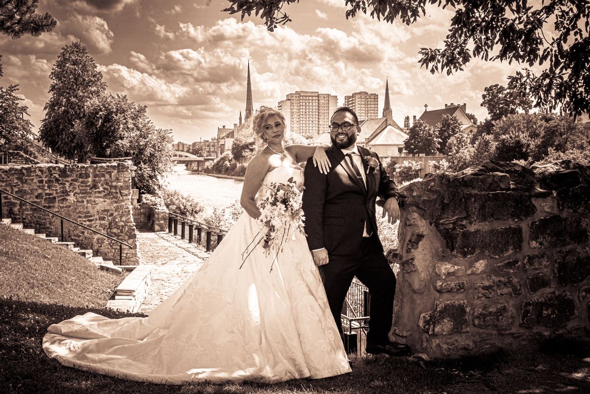 A smiling bride and groom pose beside a stone wall with historic and modern architecture in the background.