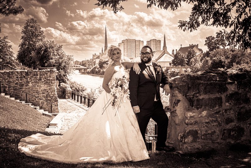 A smiling bride and groom pose beside a stone wall with historic and modern architecture in the background.