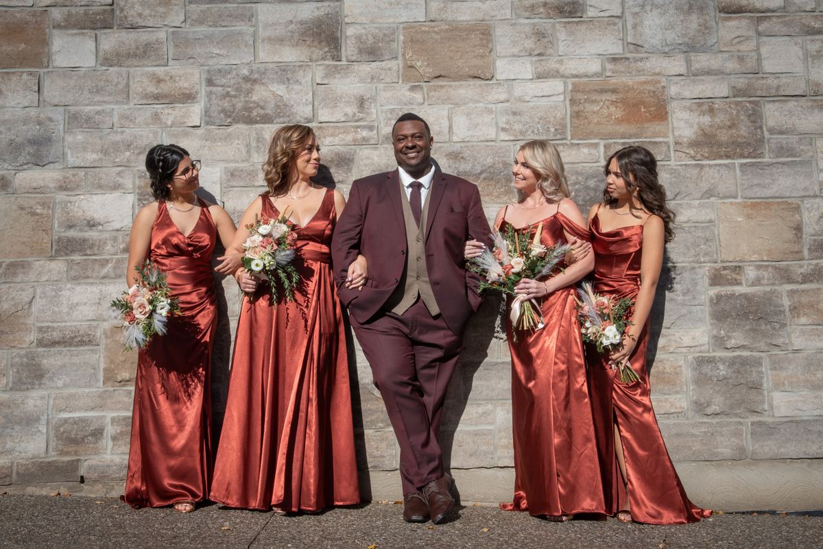 A groom in a burgundy suit stands with four bridesmaids in rust gowns, smiling against a stone wall.