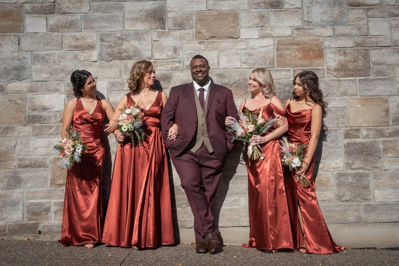 A groom in a burgundy suit stands with four bridesmaids in rust gowns, smiling against a stone wall.