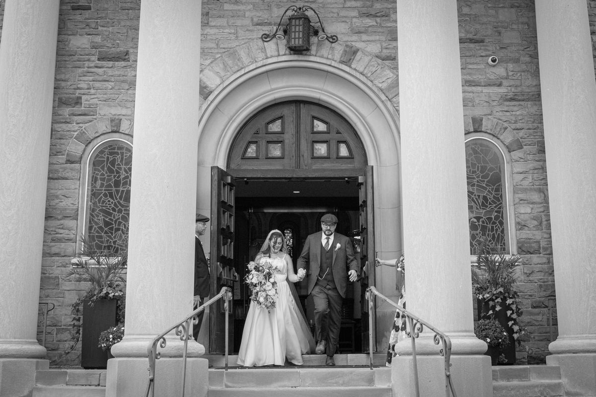 A bride and groom exit a stone church, smiling and holding hands under an arched doorway in soft black-and-white light.