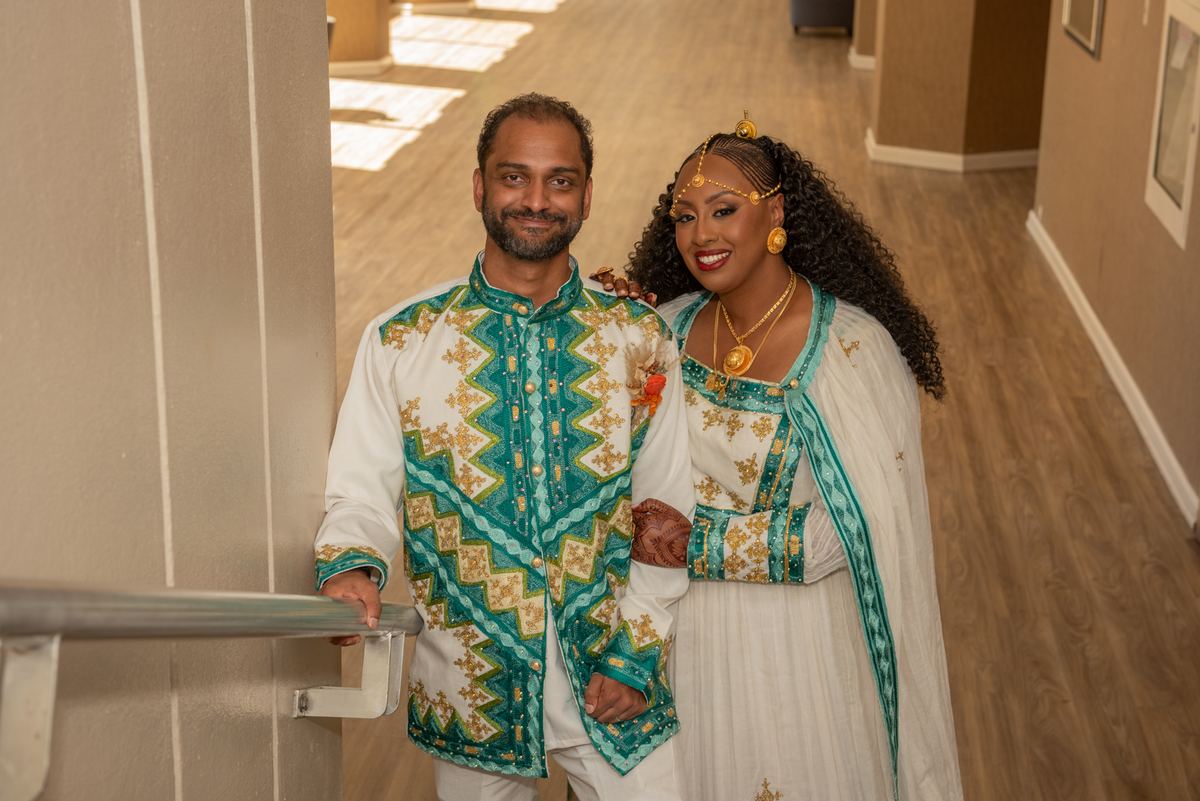 A smiling couple in ornate traditional attire with gold and teal embroidery stands by a railing, celebrating their union.