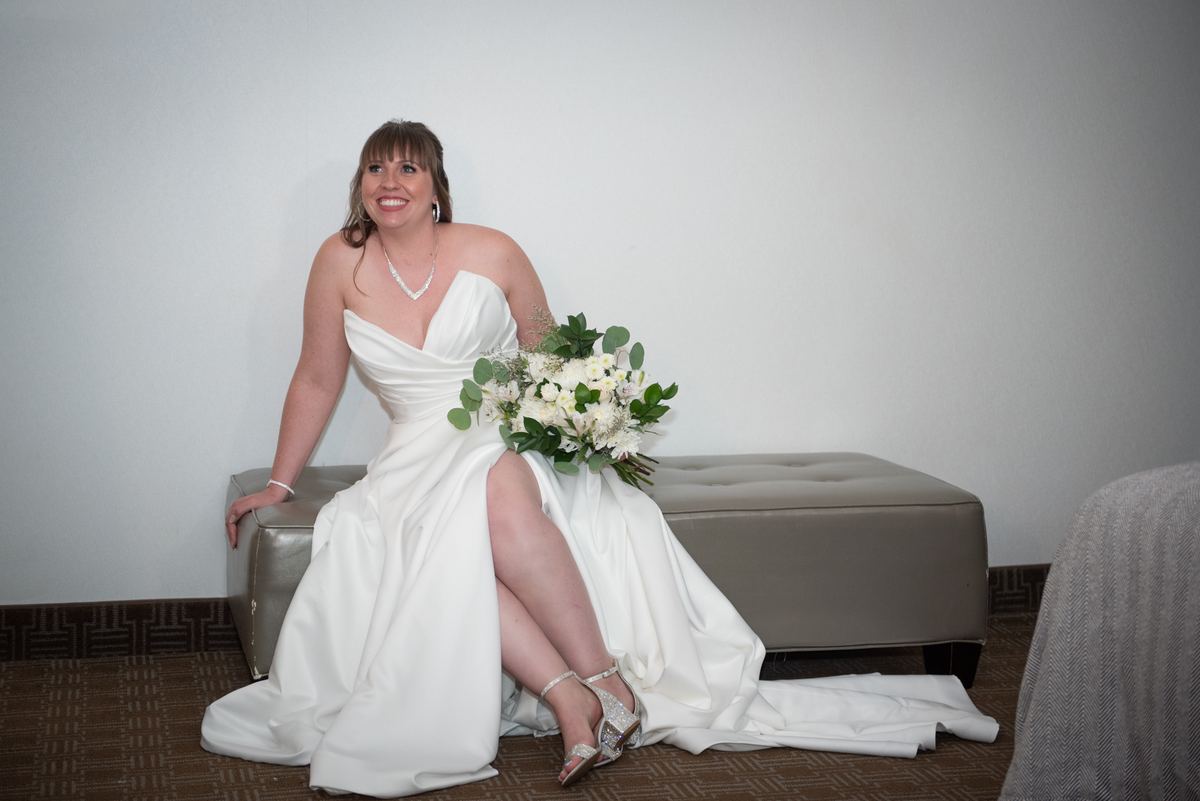 A bride sits on a modern ottoman with her leg crossed, holding a bouquet of white flowers and greenery in an indoor setting.