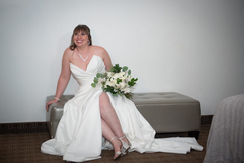 A bride sits on a modern ottoman with her leg crossed, holding a bouquet of white flowers and greenery in an indoor setting.