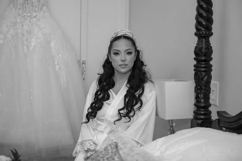 A bride in a white silk robe and tiara sits quietly, gazing at the camera near her hanging wedding gown.