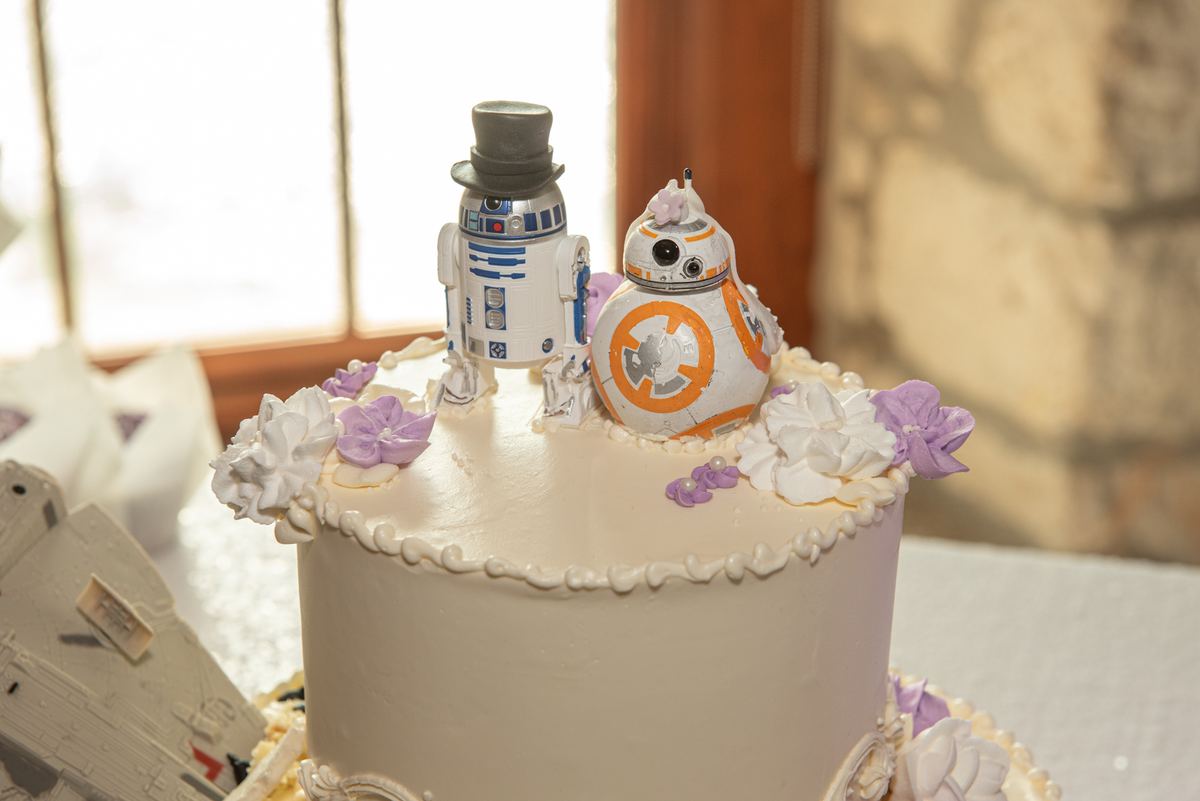 A wedding cake adorned with R2-D2 and BB-8 droids, surrounded by purple and white flowers, illuminated by soft natural light.