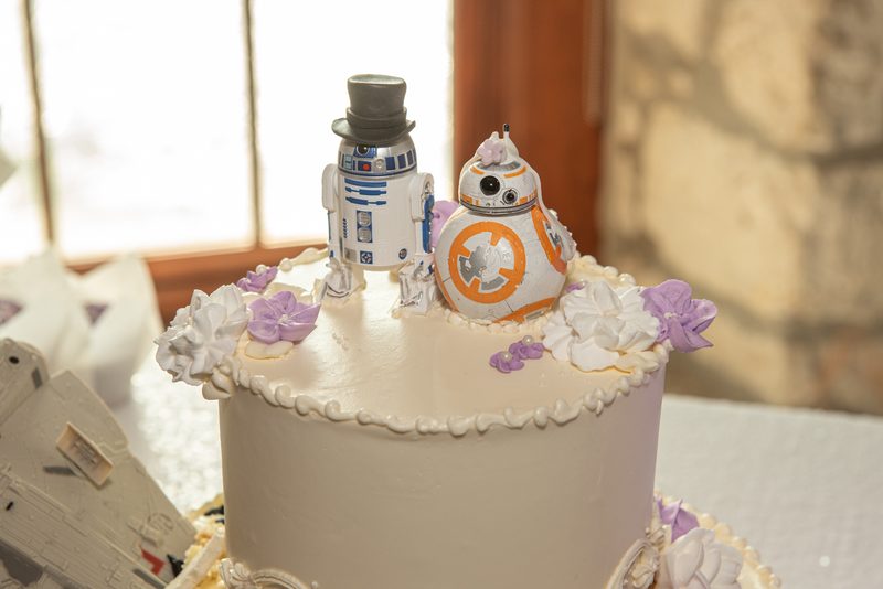 A wedding cake adorned with R2-D2 and BB-8 droids, surrounded by purple and white flowers, illuminated by soft natural light.