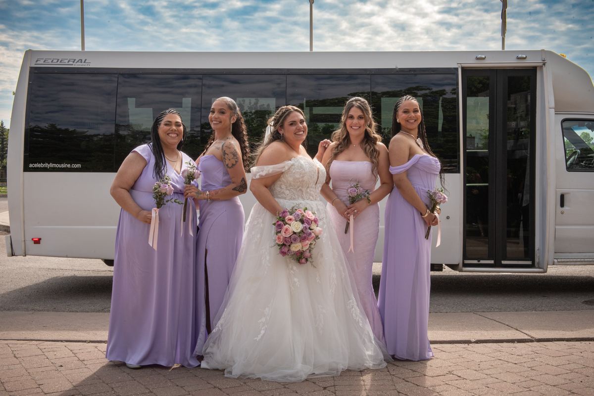 A bride in a white off-the-shoulder gown stands with four bridesmaids in lavender gowns near a limousine.