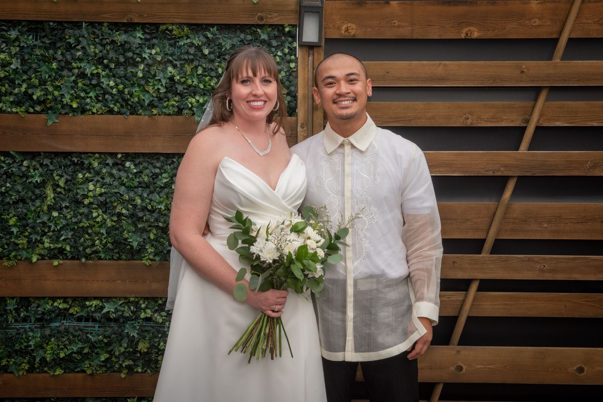 A smiling bride in a white gown stands beside her groom in a white barong tagalog against wooden slats and green ivy.