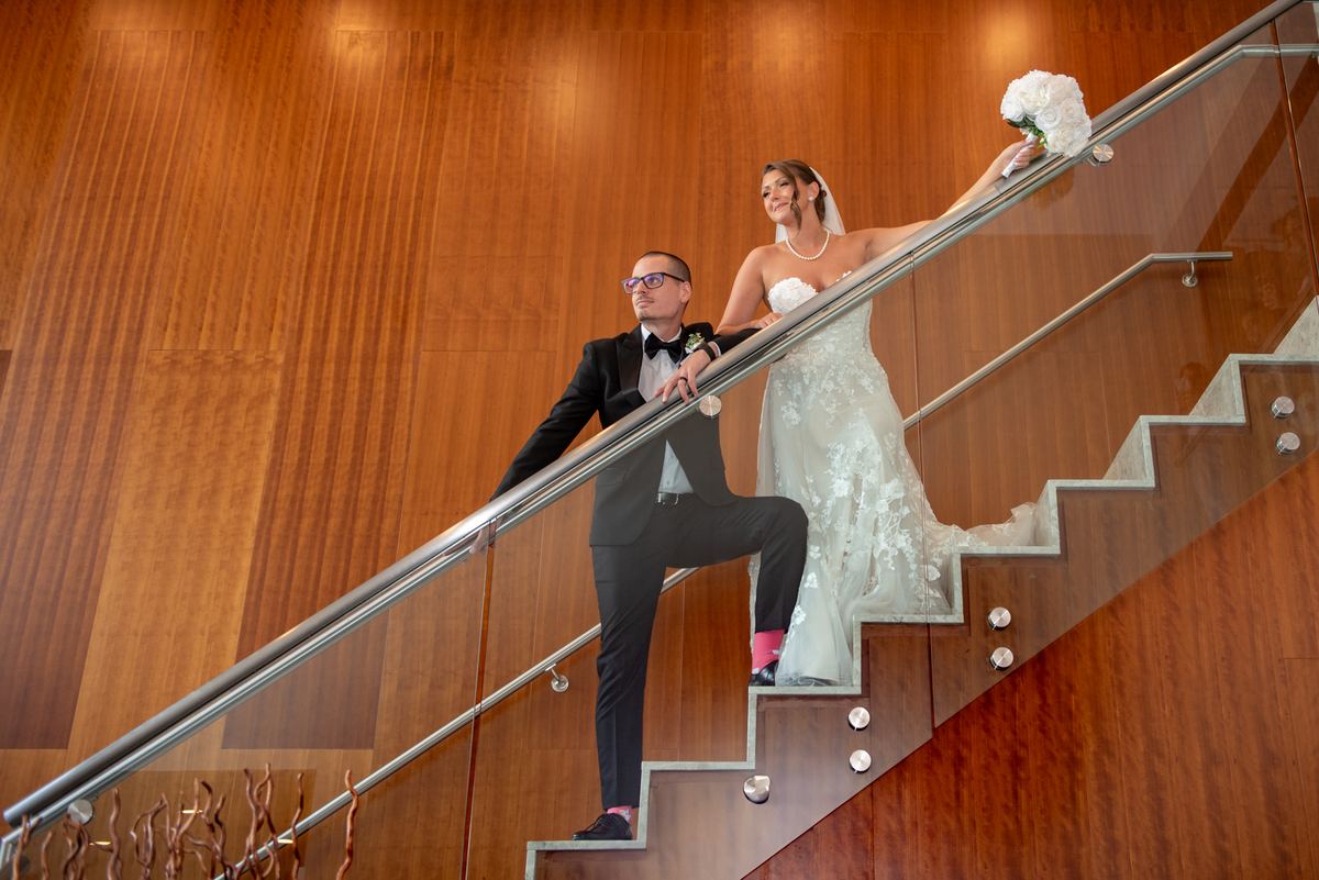A bride and groom pose playfully on a modern staircase, the bride leaning over the railing with her bouquet.