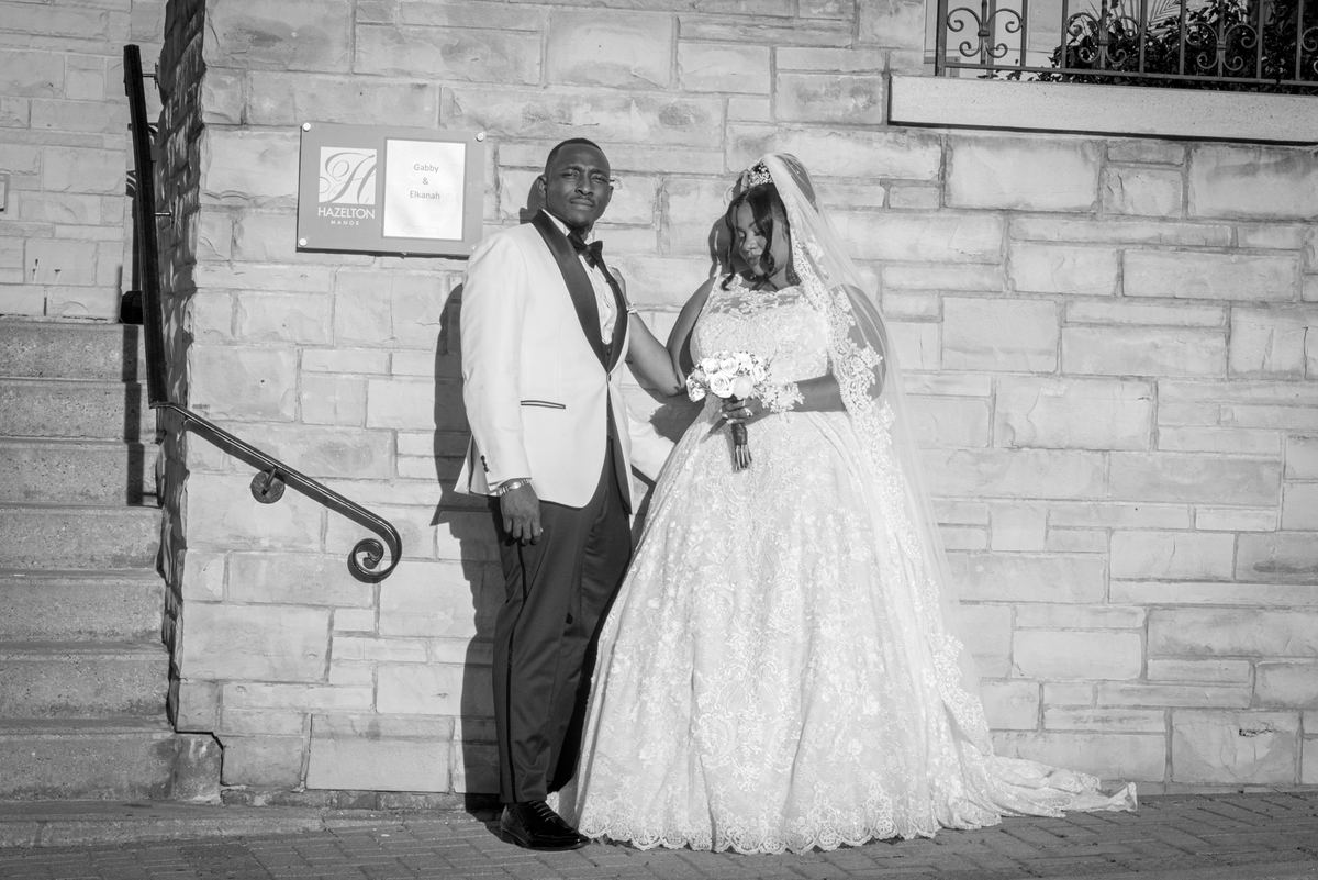 A bride in a lace gown and veil with a bouquet stands beside her groom in a tuxedo against a stone wall.