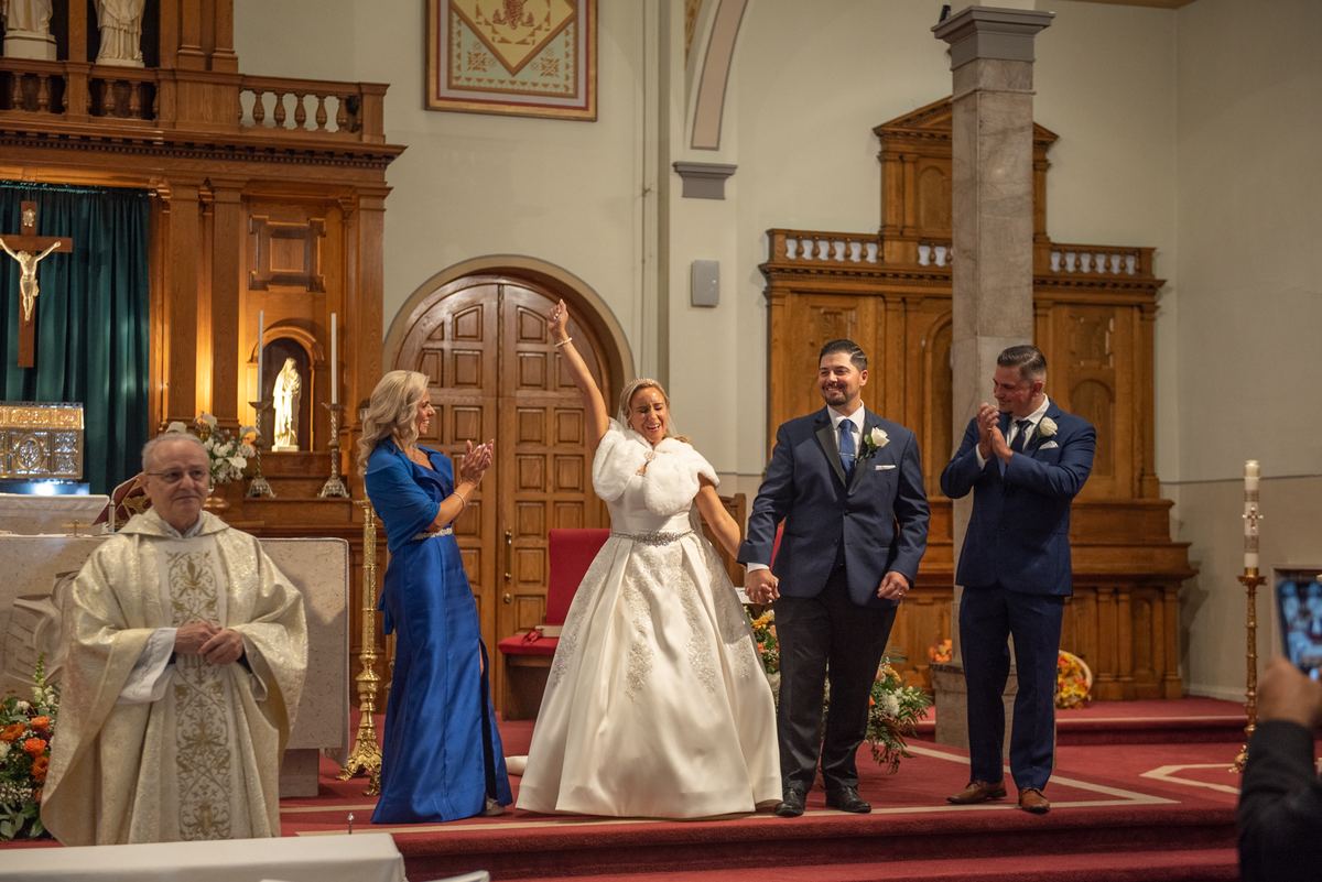 Bride and groom walk down the aisle, smiling and waving as guests clap, with ornate wooden church interior in background.