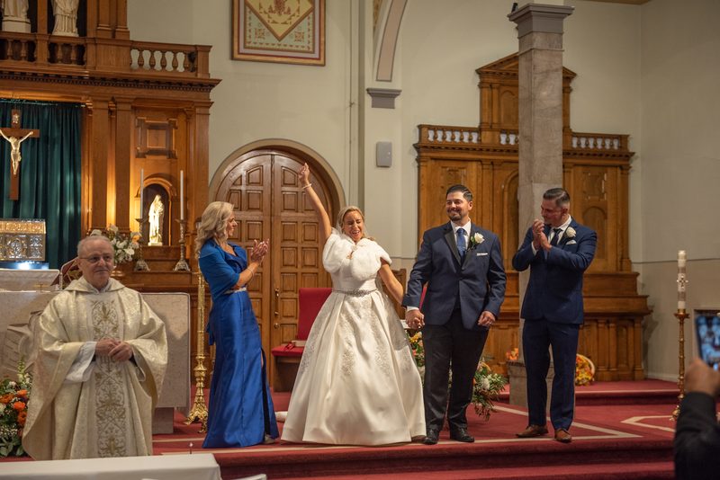 Bride and groom walk down the aisle, smiling and waving as guests clap, with ornate wooden church interior in background.
