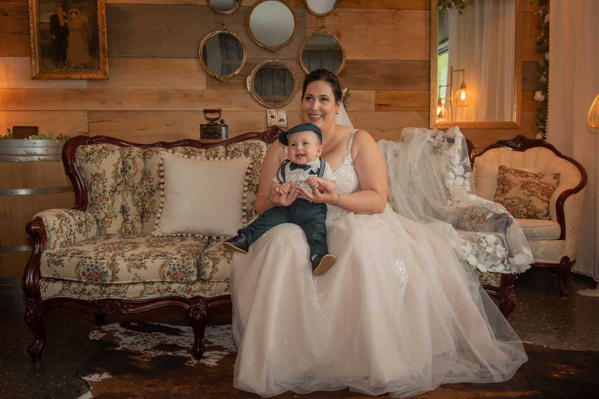 A bride in a white gown sits with a baby dressed in a tiny bowtie and cap on an ornate floral sofa in a rustic room.