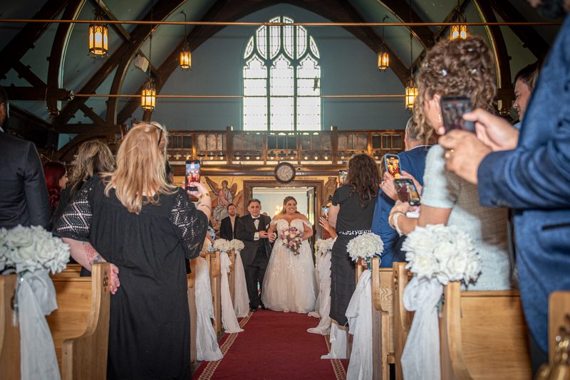 A bride in white walks down the aisle of a wooden church, illuminated by stained glass and lanterns.
