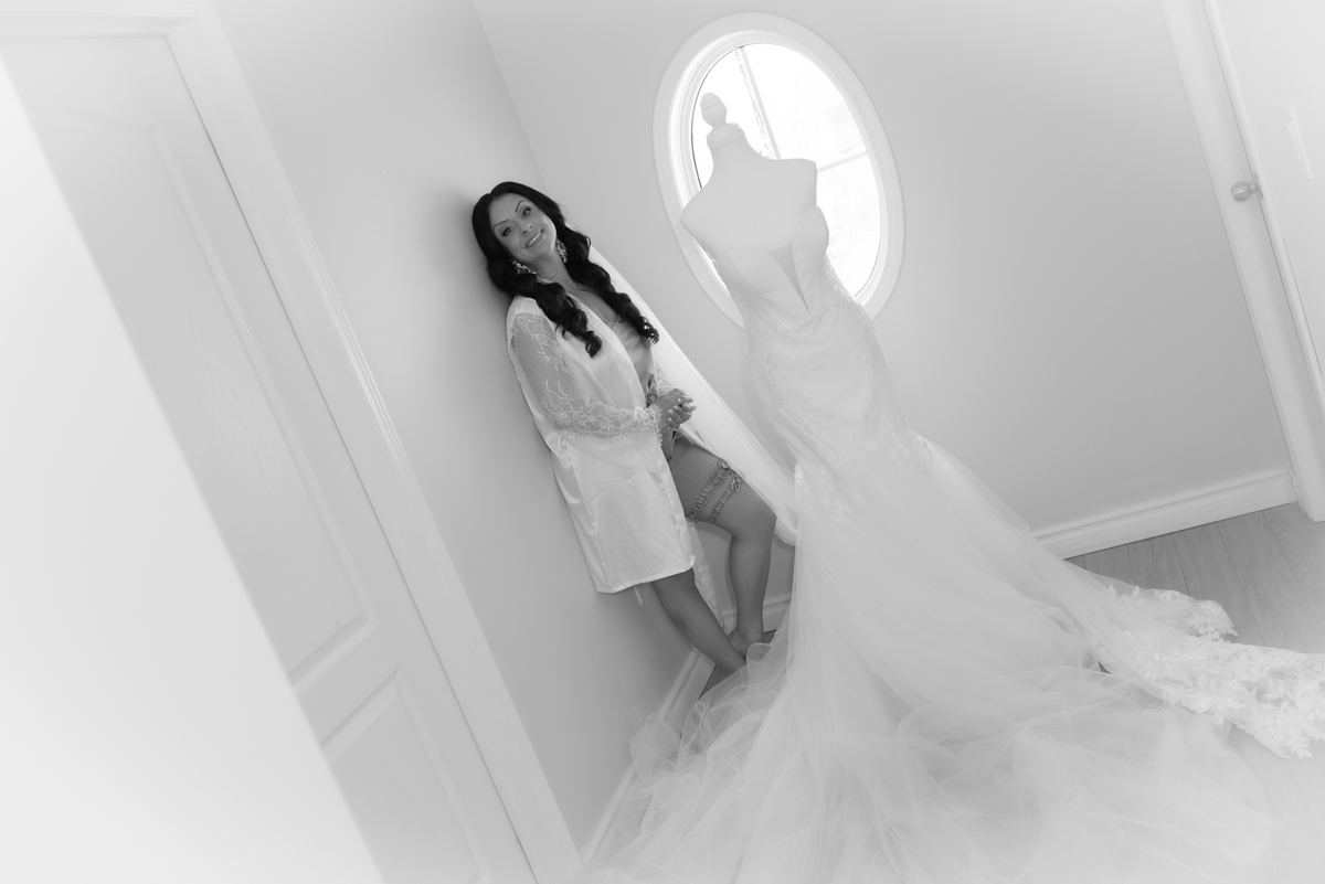 A bride-to-be leans against a wall beside a mannequin draped in her full-length wedding gown, smiling softly.