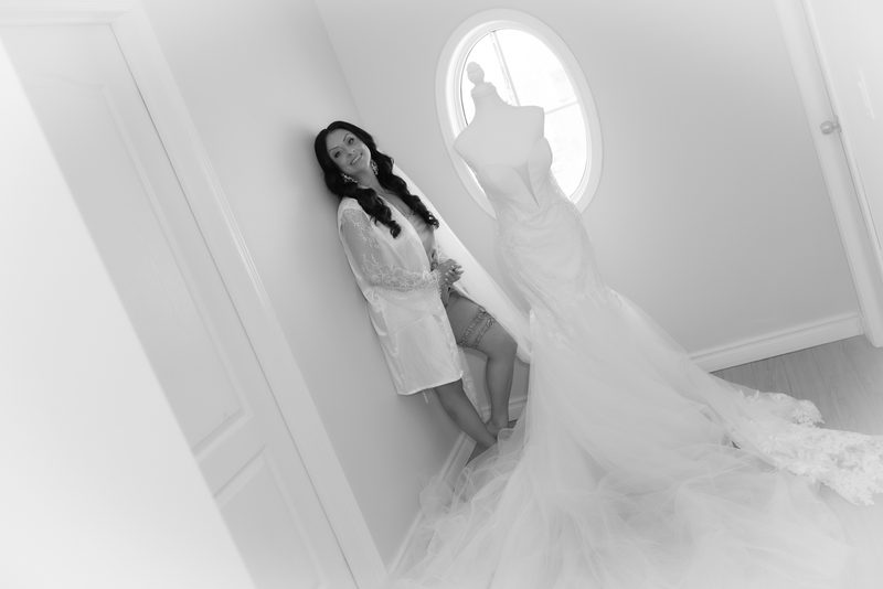 A bride-to-be leans against a wall beside a mannequin draped in her full-length wedding gown, smiling softly.