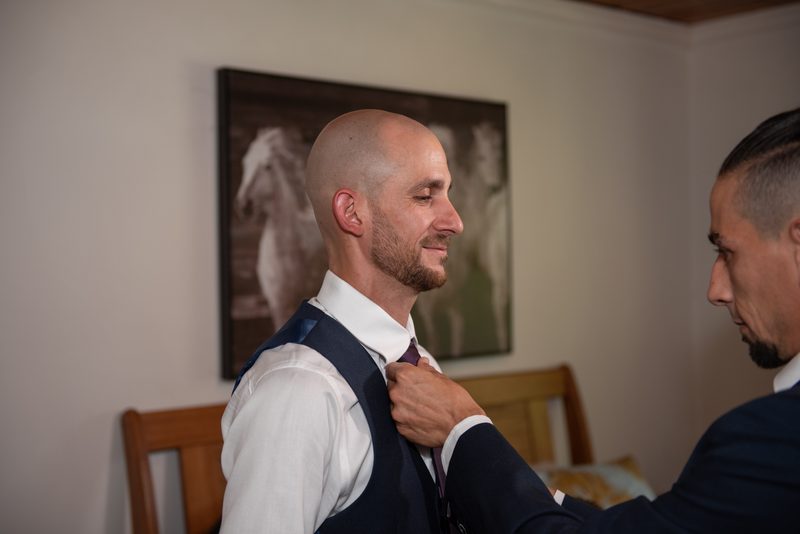 A groom and a friend prepare for the ceremony, adjusting ties in an intimate room with wooden furniture.