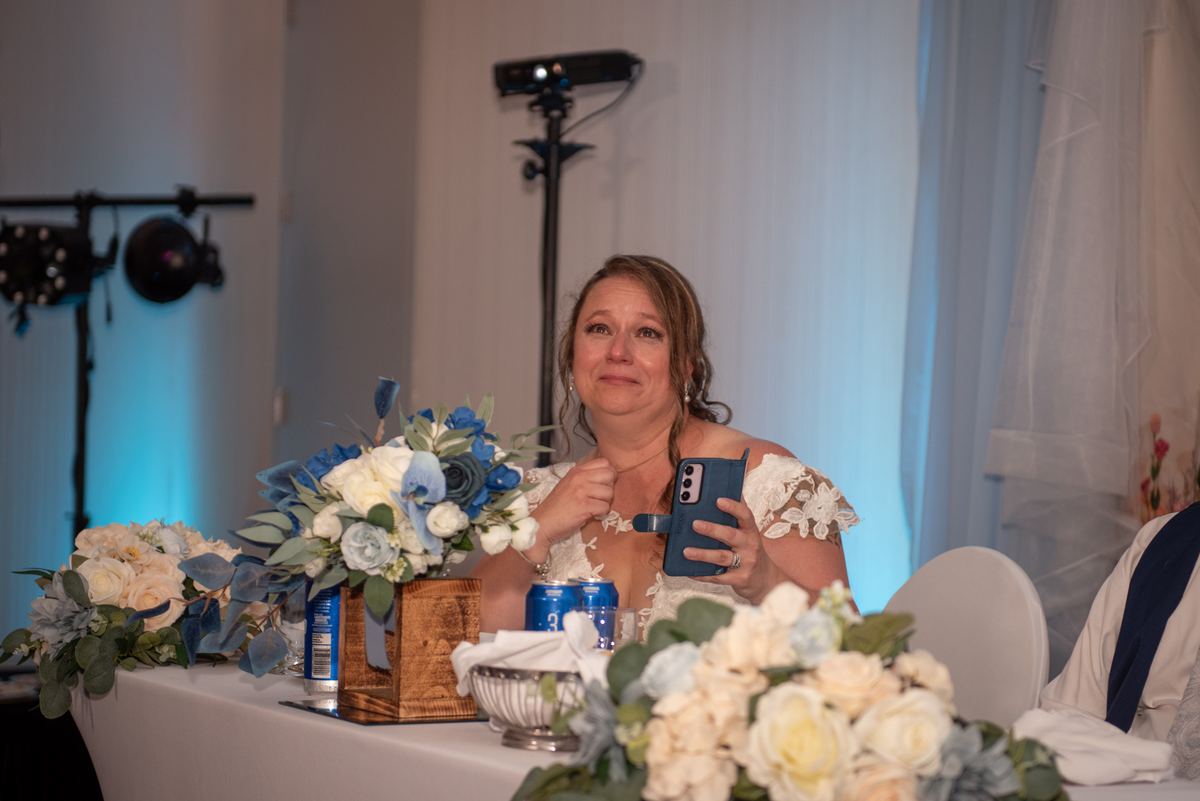 A woman at a decorated table holds her phone, watching an emotional ceremony with blue lighting and floral arrangements.