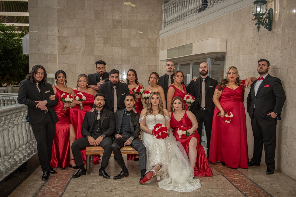 A bride in white sits with her groom and wedding party in formal attire holding red-and-white flowers.