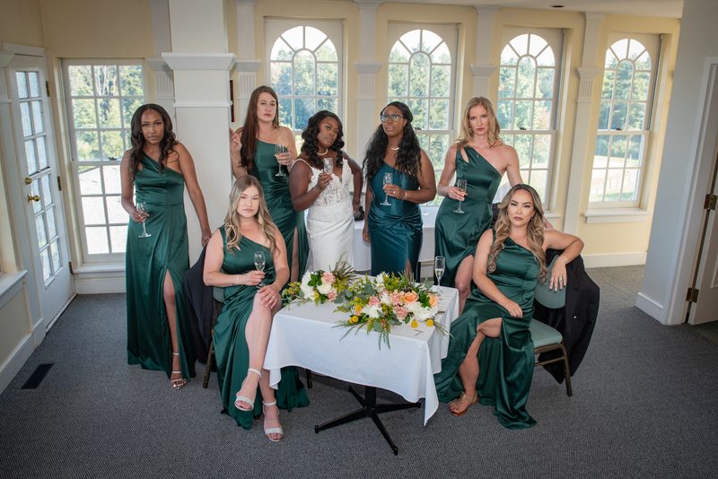 A bride and her bridesmaids in emerald green dresses hold champagne flutes near large windows, illuminated by natural light.