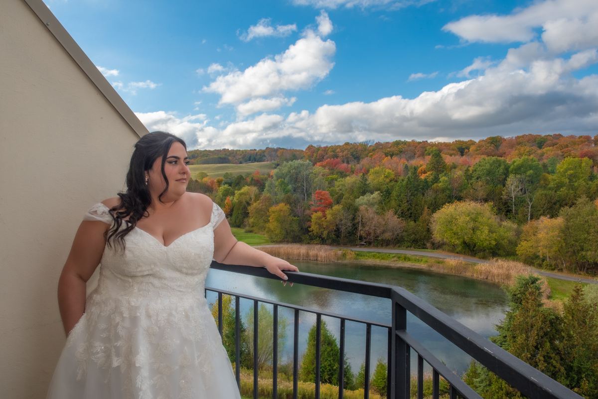 A bride in a white lace gown stands pensively on a balcony overlooking an autumn landscape and lake.