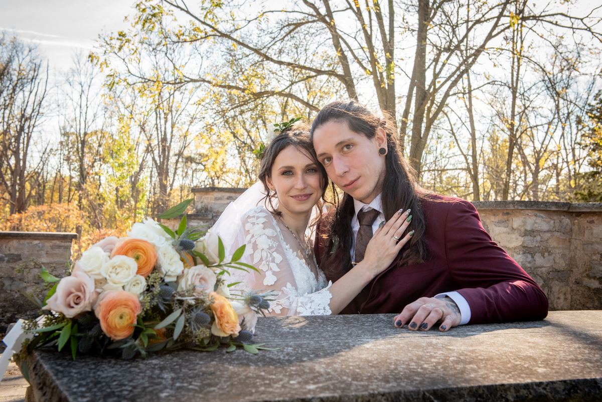 A bride in a lace gown leans on her groom beside a stone bench, surrounded by autumn leaves and flowers.