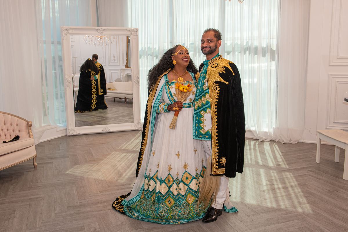 A smiling couple in ornate traditional attire stands in an elegantly appointed room with sunlight streaming through curtains.