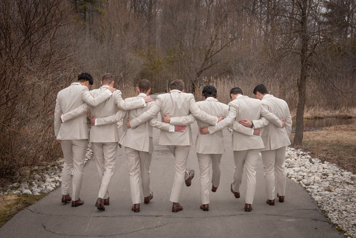 Groomsmen in beige suits walk arm-in-arm down a paved path in a quiet, wooded area on an overcast day.