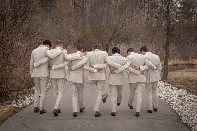 Groomsmen in beige suits walk arm-in-arm down a paved path in a quiet, wooded area on an overcast day.