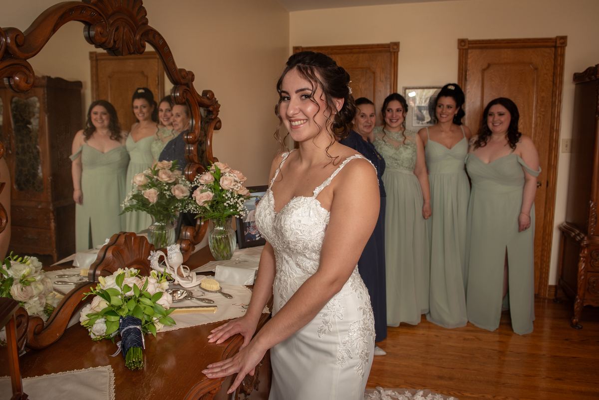 A bride in a white lace gown smiles by a wooden dresser with her sage green-clad bridesmaids near a carved mirror.